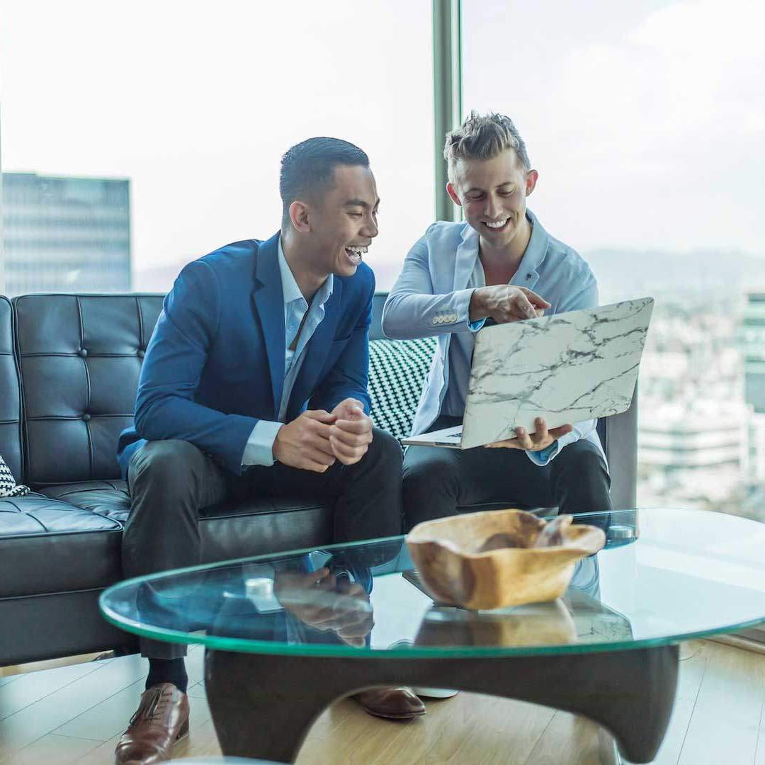 Two smiling men sitting on an office couch looking at an open laptop